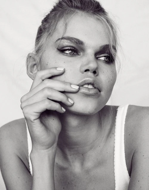 Close-up black and white portrait of a young woman with smokey eyes, her hand near her face, against a bright neutral background.