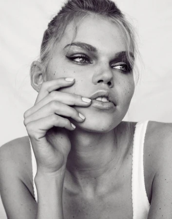 Close-up black and white portrait of a young woman with smokey eyes, her hand near her face, against a bright neutral background.