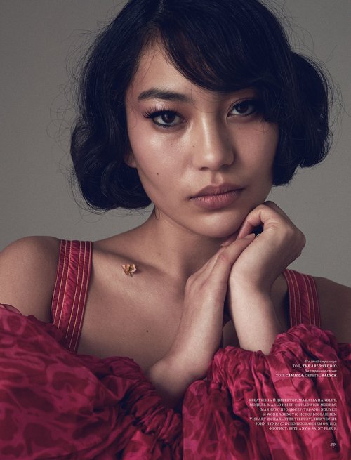 Close-up portrait of a young Asian woman with dark wavy hair, wearing a red patterned off-the-shoulder top in a photo studio setting.