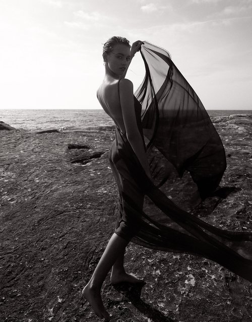 Monochrome portrait of a woman in a sheer dress holding fabric, standing on rocks near the ocean under a bright sky. Fashion.