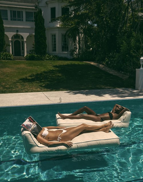 Two women in bikinis relax on floating loungers in a pool outside a large white house with a well-manicured green lawn.