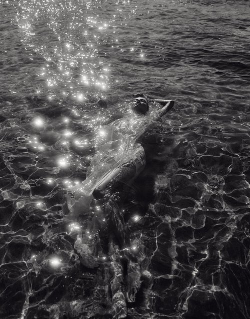 Black and white image of an adult woman floating in rippling water with sunlight reflecting off the surface. She is wearing a light dress.