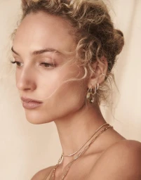 Close-up portrait of a woman wearing gold hoop earrings and layered gold necklaces against a textured neutral background.