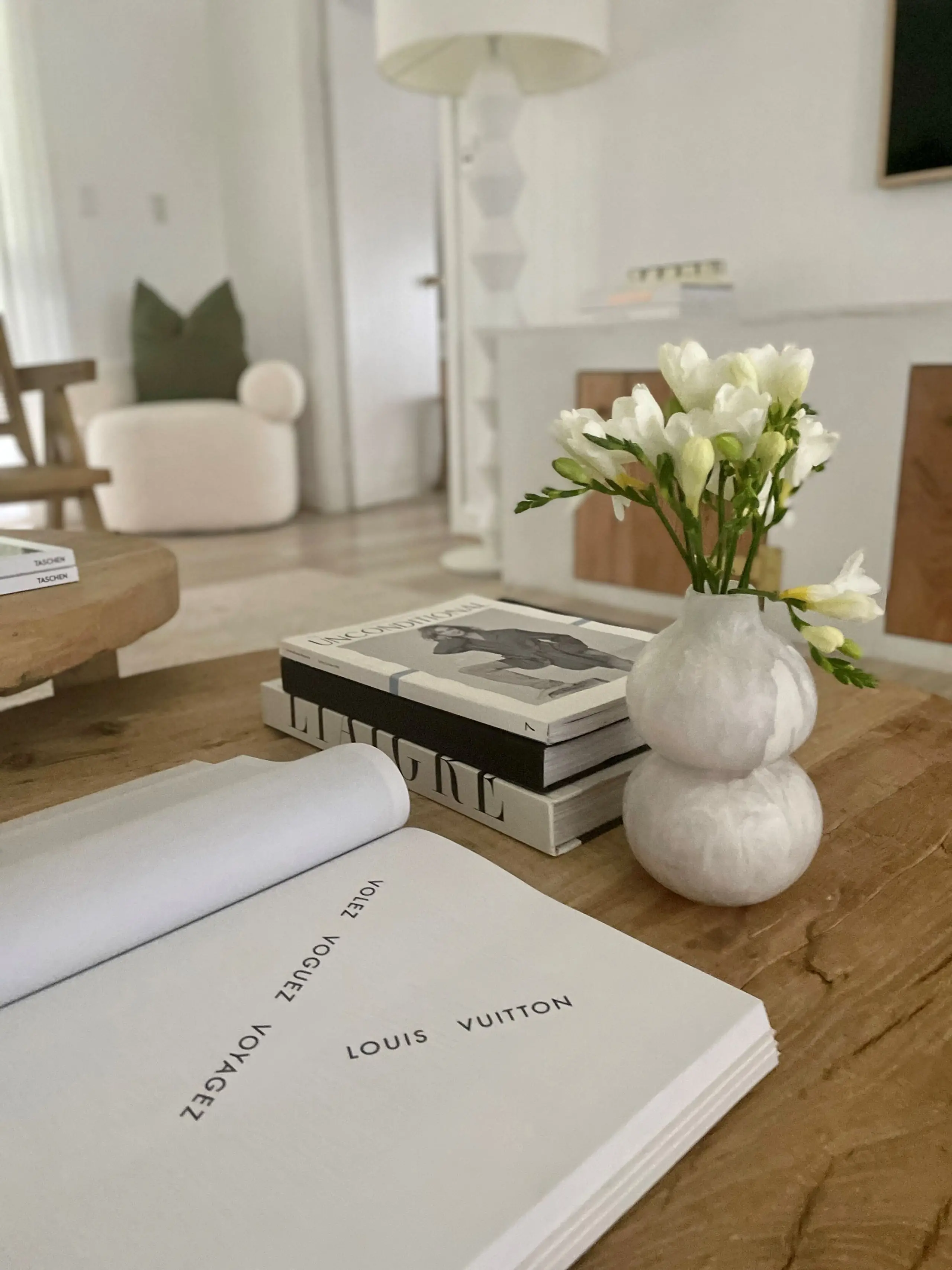 Open book with 'Louis Vuitton' text, flowers in a white vase, and fashion books on a wooden coffee table in a bright living room.