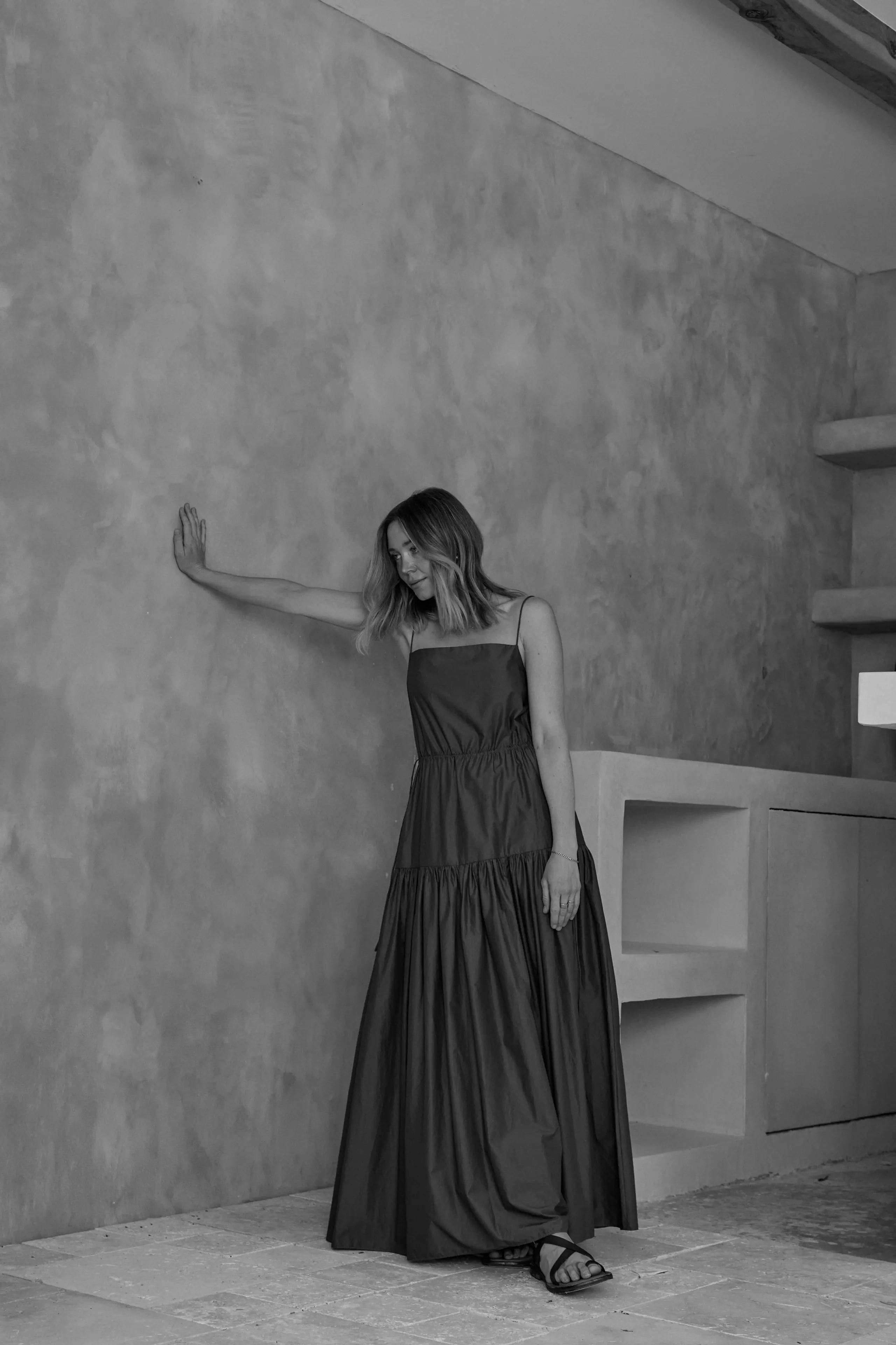 Adult woman poses in long dress indoors. The background features a textured wall. The photography is in black and white, with soft lighting.