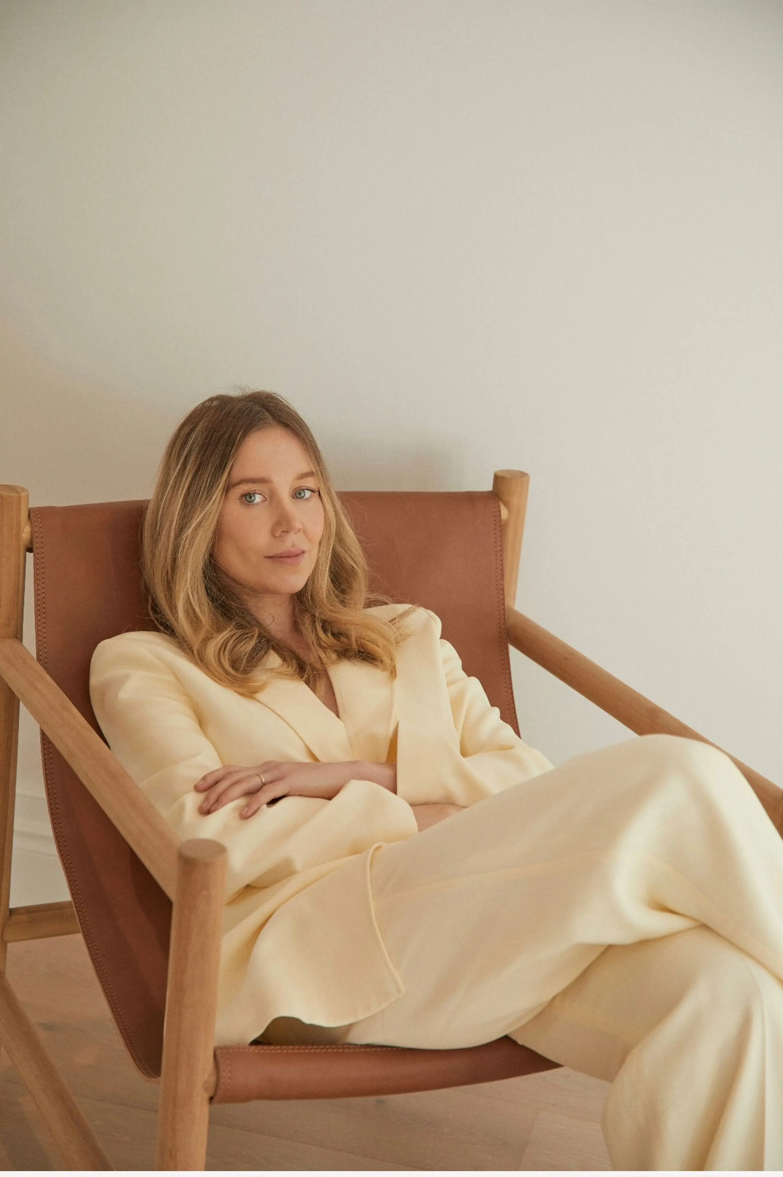 Woman in a light yellow pantsuit sits in a wood-frame chair against an off-white wall in a relaxed pose with arms crossed.
