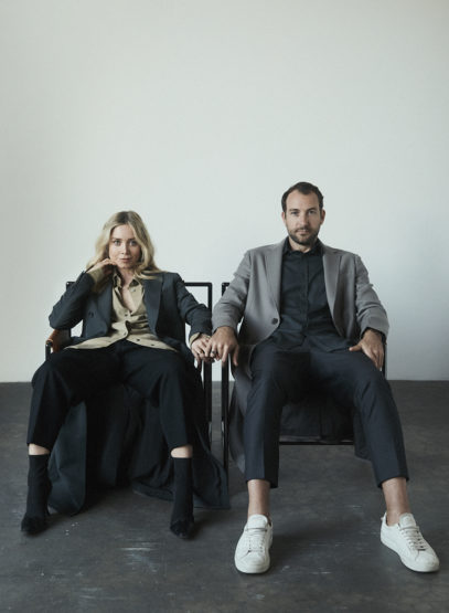 A stylish couple sitting side-by-side in a studio, wearing fashionable attire and holding hands against a stark white background.