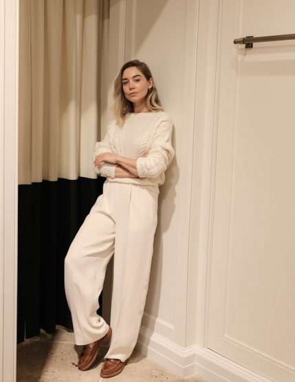 Blonde woman in cream sweater and slacks leans against a white wall in brown loafers, with a neutral-toned curtain backdrop.