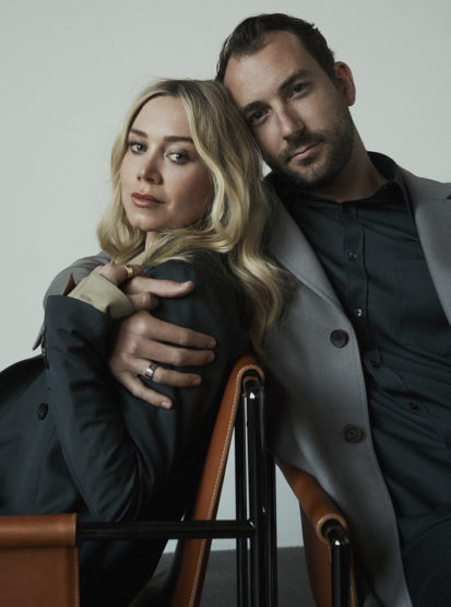 Portrait of a couple in studio, the blonde woman sits on a chair while the dark-haired man stands behind with his hand on her shoulder.