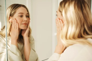 Blonde woman looking in a bathroom mirror holding her face with both hands styled by Hayley Bonham
