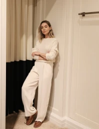 Woman in cream knit sweater and white trousers leaning against a wall, styled by Hayley Bonham in Sydney.