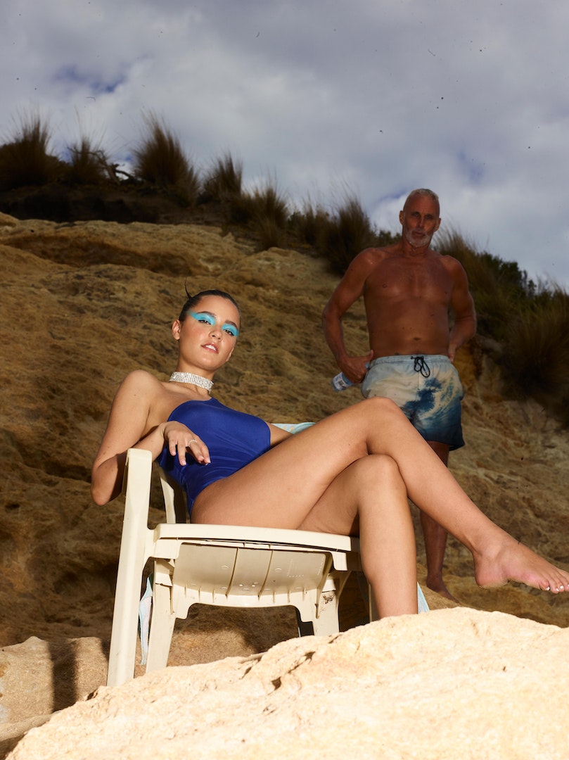Woman in blue swimsuit poses on a white plastic chair near a shirtless adult male on a beach with a sandy cliff and cloudy sky.
