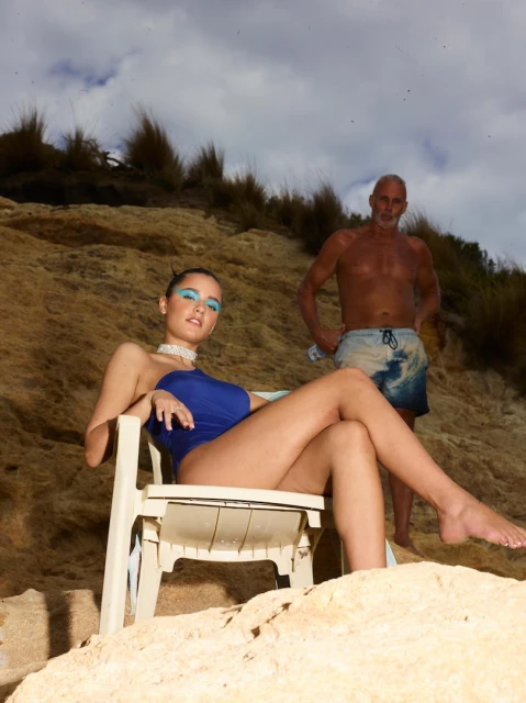 Woman in blue swimsuit poses on a white plastic chair near a shirtless adult male on a beach with a sandy cliff and cloudy sky.