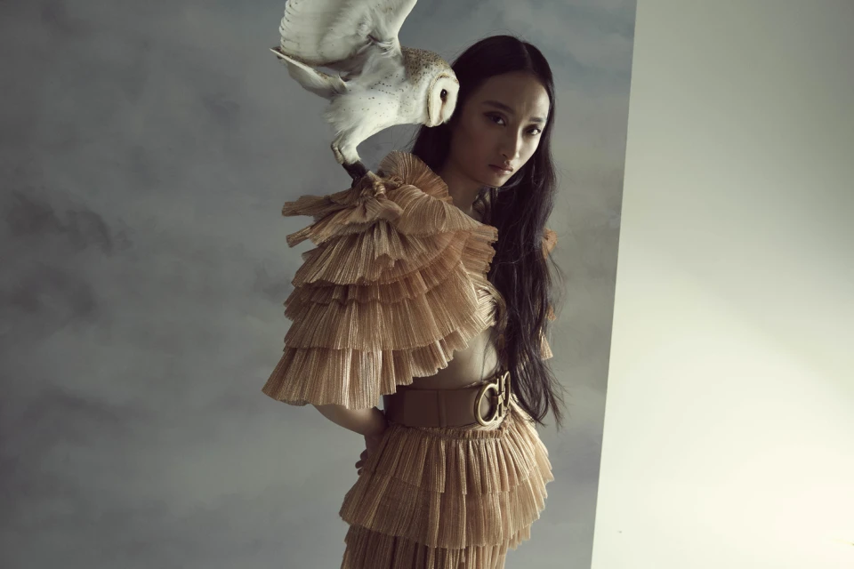 Artistic fashion portrait of Asian woman in tiered tan dress with white owl perched on shoulder against a muted grey background.