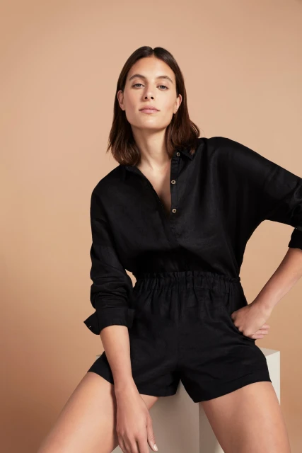 Portrait of a woman wearing a black collared shirt and shorts, posing indoors with her hand in her pocket in a studio setting.