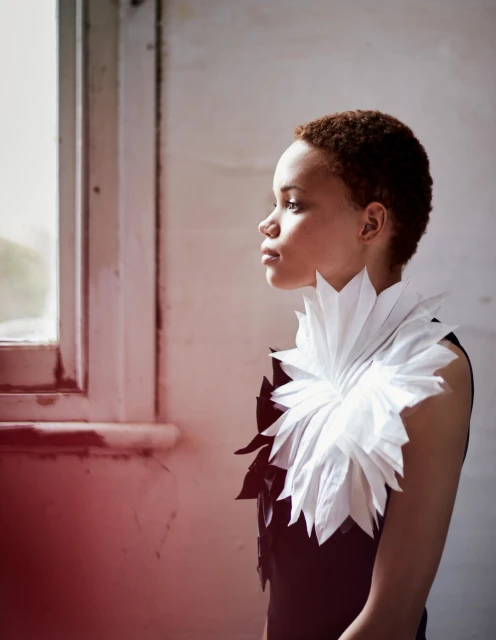 Woman in sculptural white and black paper dress poses near a window in a minimalist interior, illuminated by soft natural light.