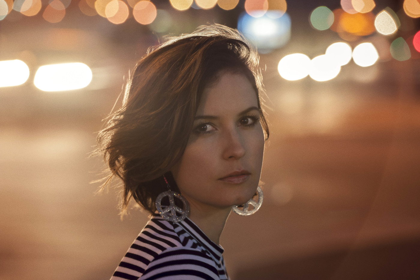 A young adult woman with short brown hair poses in front of blurred lights while wearing a striped shirt and peace sign earrings.
