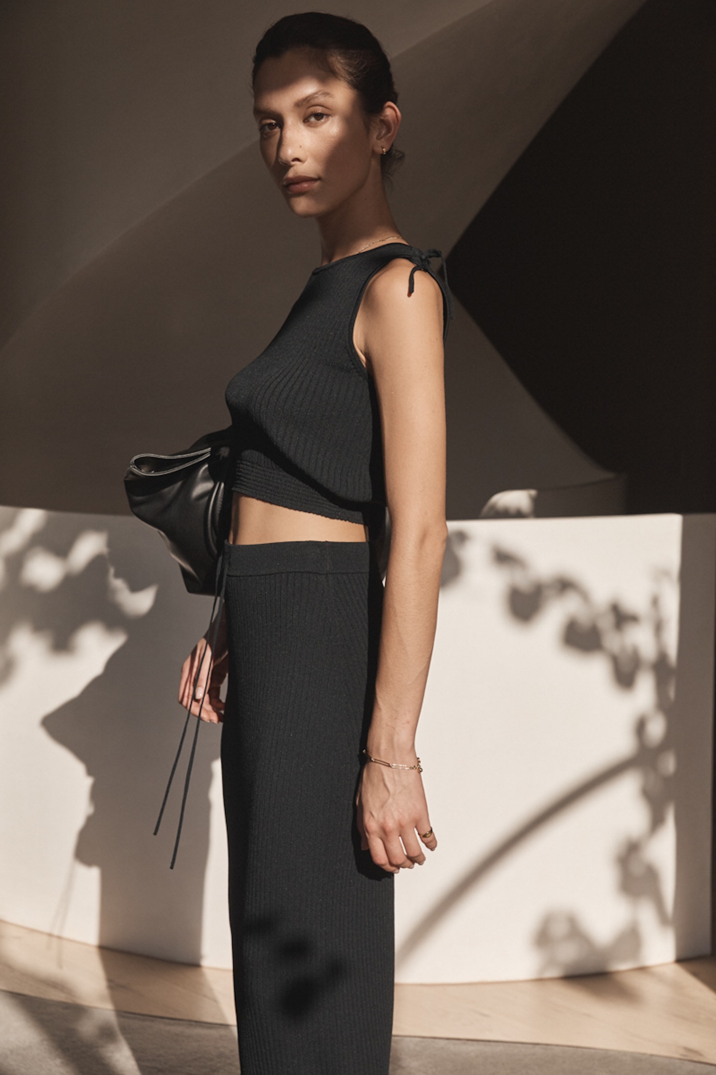 An adult female model poses indoors wearing a black knit top and skirt, accessorized with a black leather bag and bracelet.