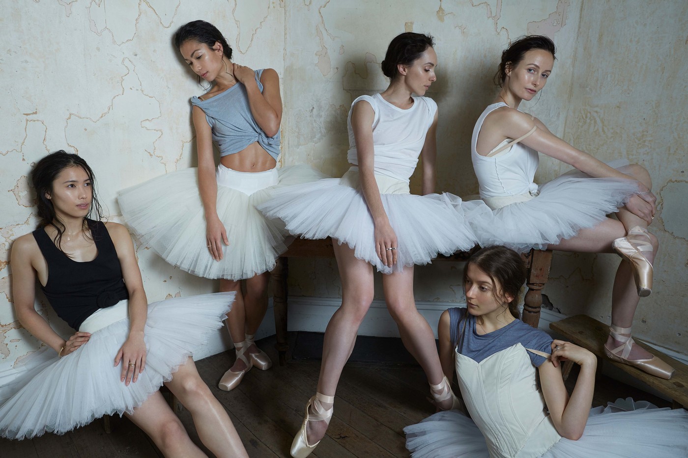Five ballet dancers in leotards and tutus strike poses in a room with distressed walls and a hardwood floor, bathed in natural light.