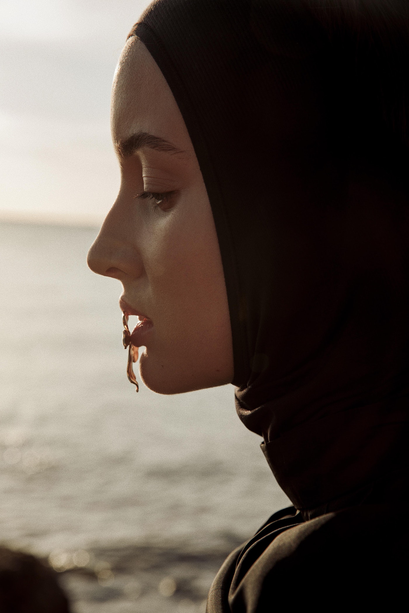 Portrait of a woman with a headscarf, shot in profile with a blurred ocean and sky background in soft, natural lighting. Close-up on face.