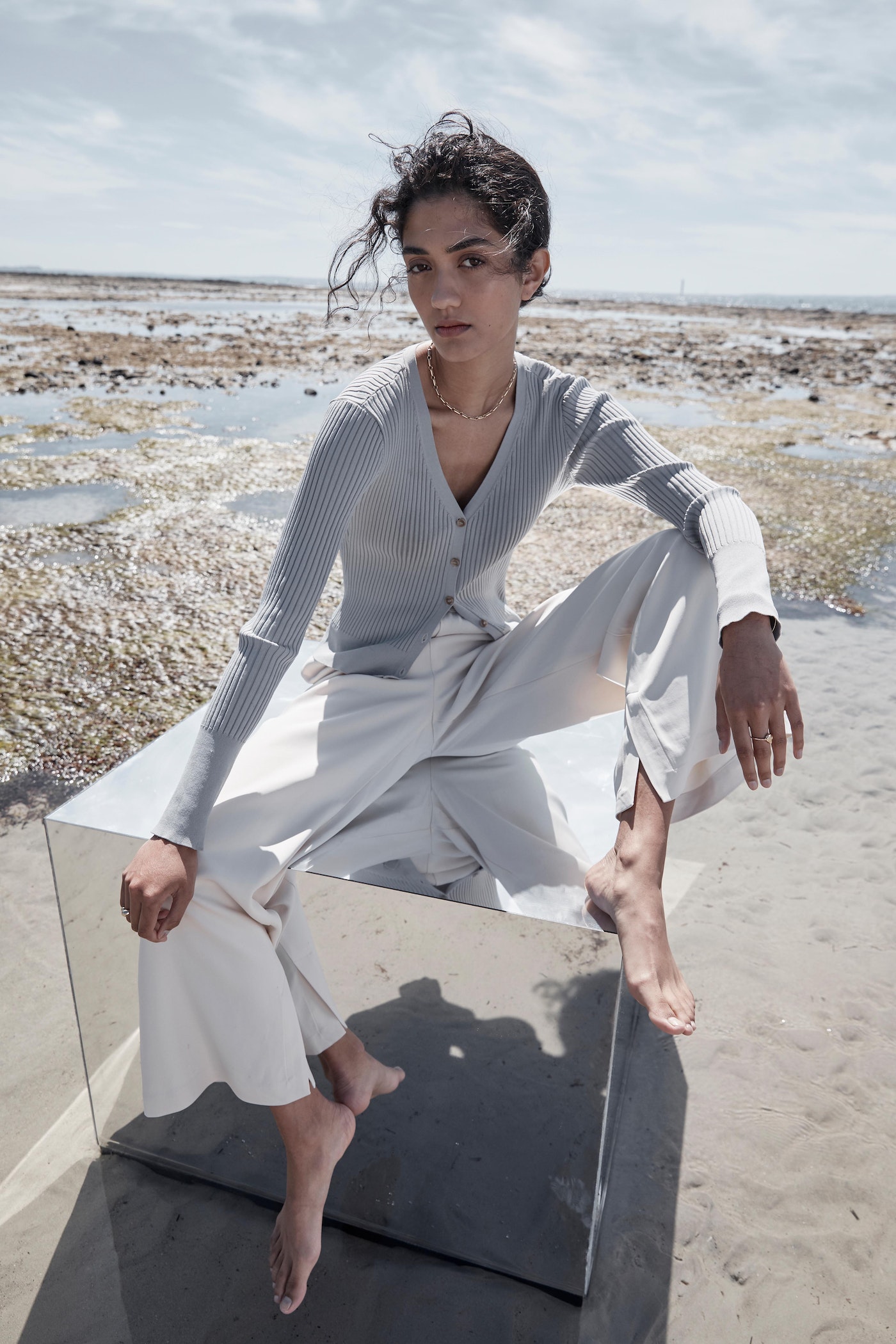 Woman in cardigan and wide-leg pants sits on a reflective cube at the beach with the ocean and sky as background.