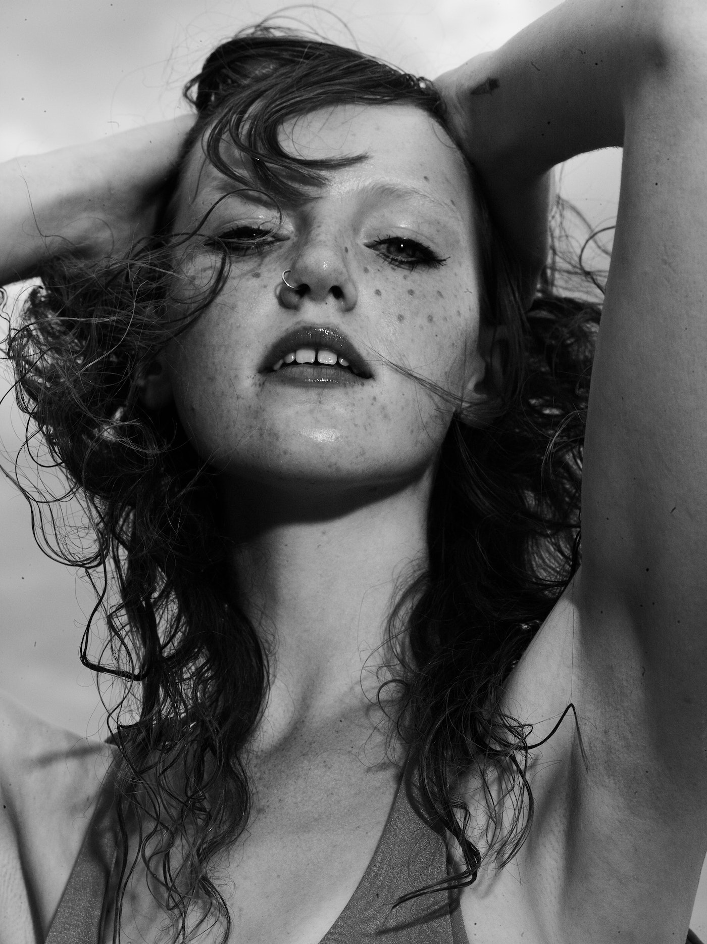 Monochrome portrait of a young woman with freckles, wet wavy hair, and a nose ring, posing with hands behind her head against a light sky.