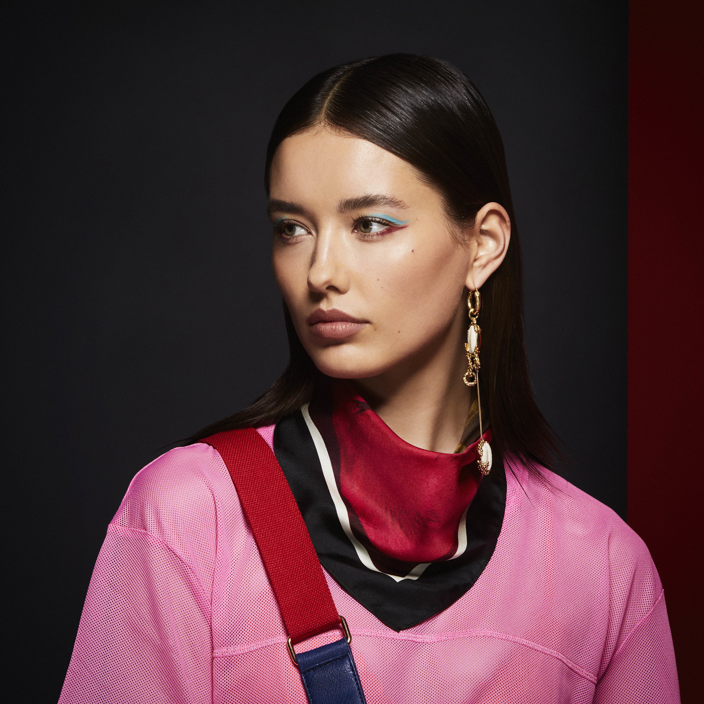 Portrait of a young woman with dark hair, wearing a pink shirt, black scarf, and statement earrings against a dark backdrop.
