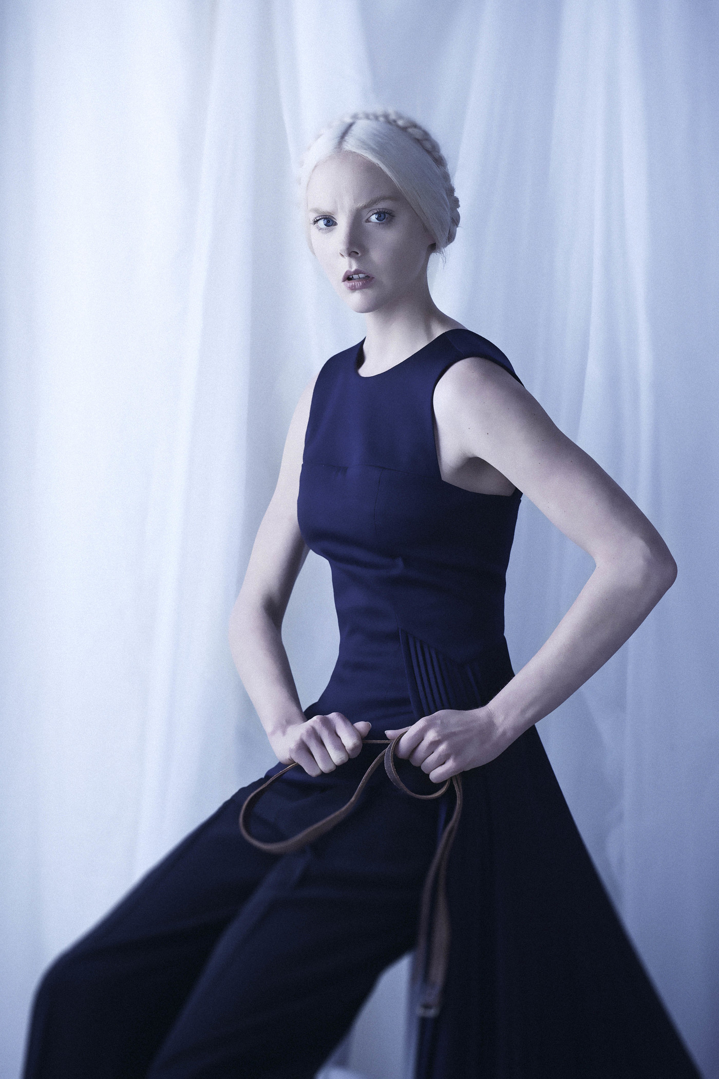 Blonde woman in a black dress poses against a light blue backdrop, holding a belt in her hands, creating a minimalist fashion portrait.