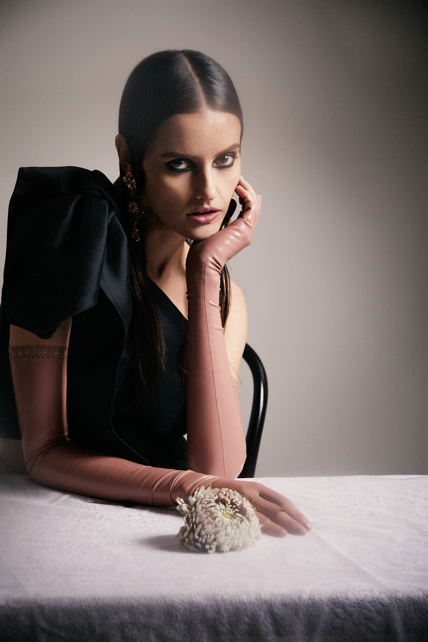 Brunette woman with gloved hand on cheek poses at table in black dress with floral prop in a studio setting, eye-level portrait.