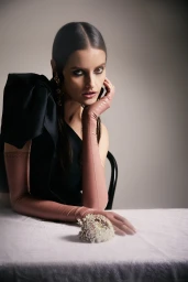 Woman with dark hair in black top and long pink gloves resting chin on hand with flower on table.
