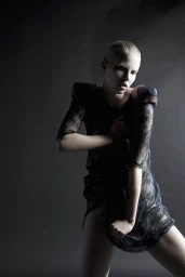 Model with slicked-back blonde hair wearing a sheer dark lace dress against a grey background