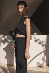 Model with slicked-back hair wearing a black ribbed two-piece outfit and holding a dark handbag against a light architectural wall.
