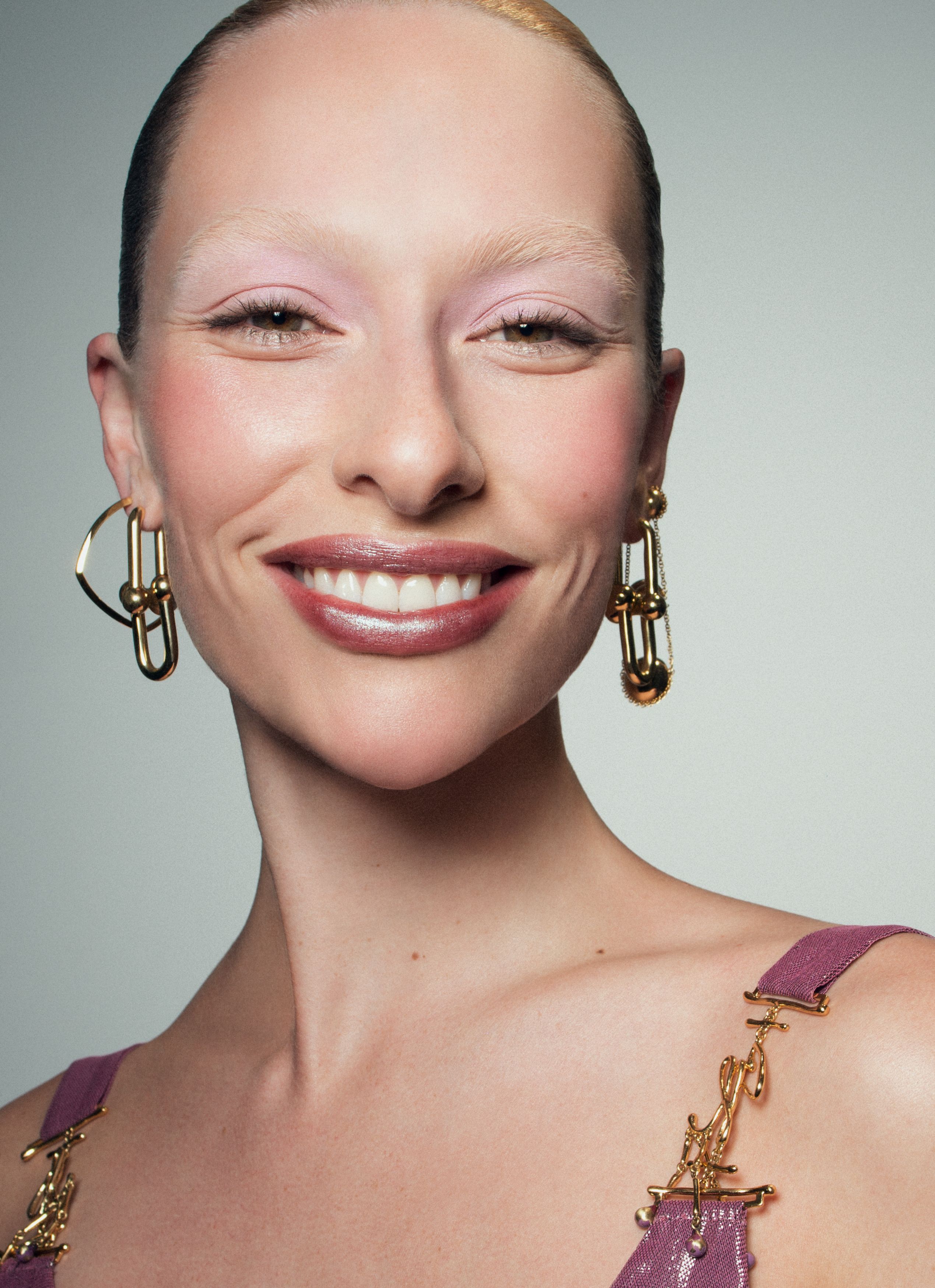 Close-up shot of a smiling woman with pale skin, light eyebrows and glossy lips, wearing gold earrings and a lavender top.
