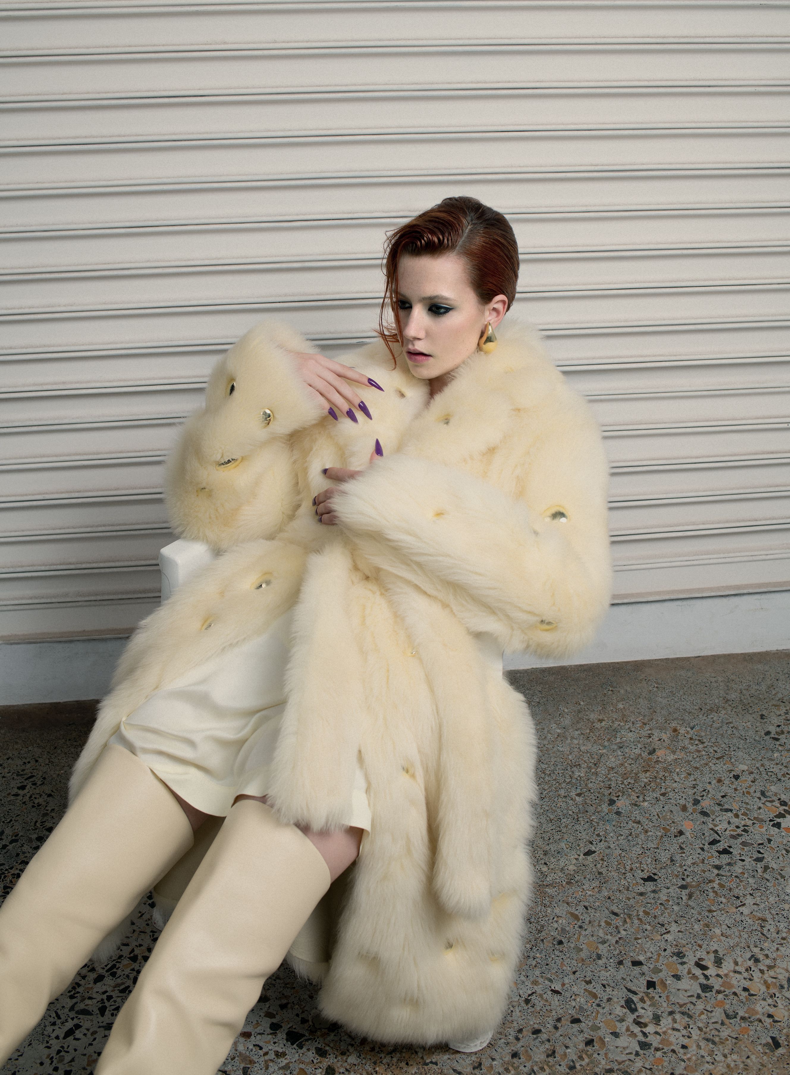 Woman with red hair in a cream-colored, knee-length dress, thigh-high boots, and a long fur coat poses in an industrial setting.