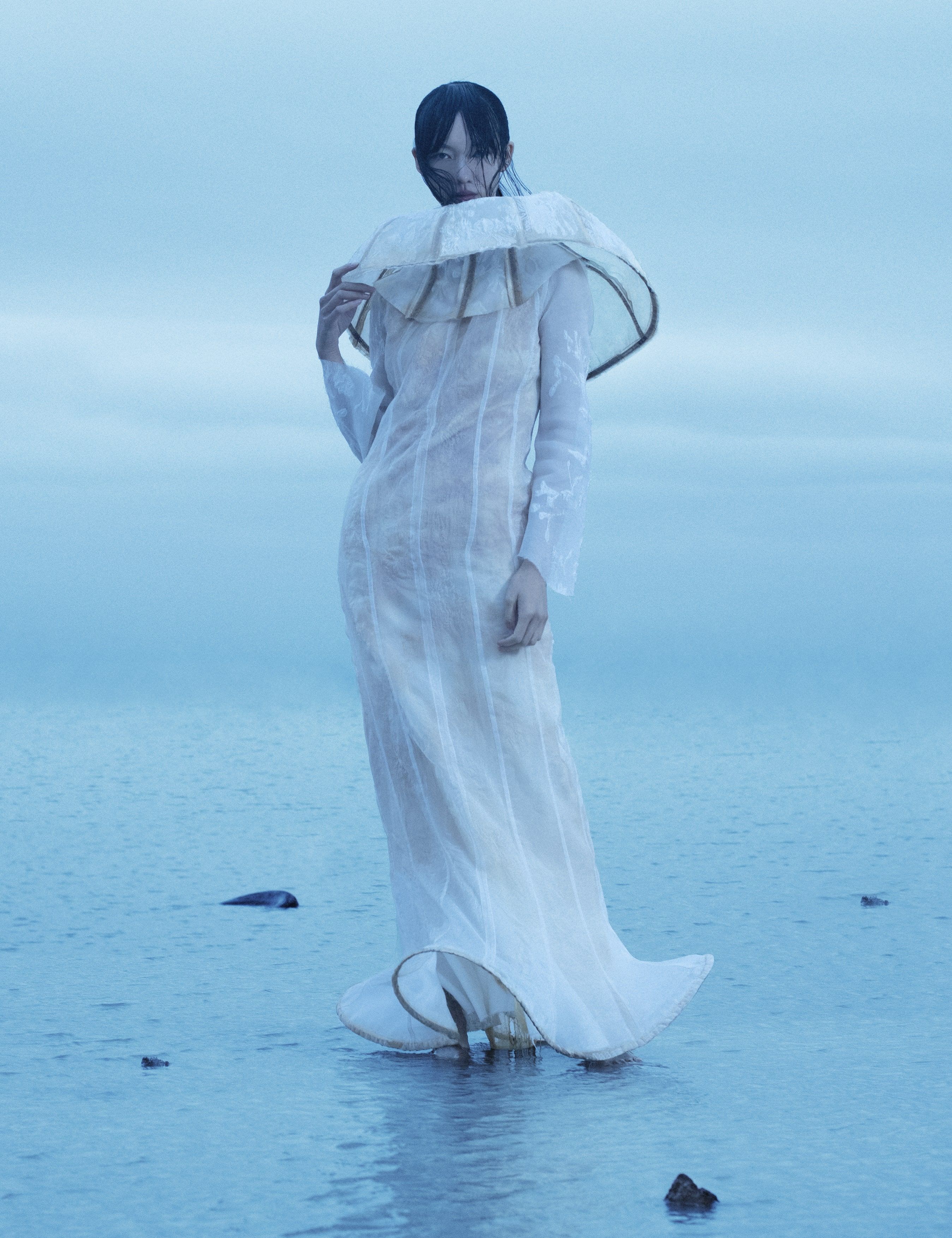 Woman stands in shallow water wearing an elaborate white dress with a large collar. Background features a soft, overcast sky.