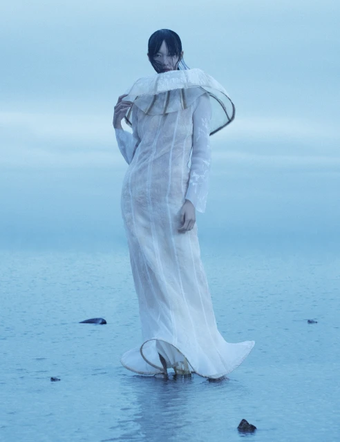 Woman stands in shallow water wearing an elaborate white dress with a large collar. Background features a soft, overcast sky.