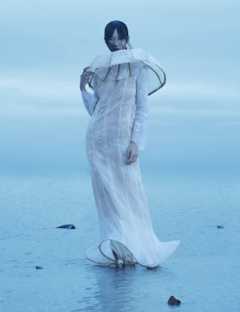Woman stands in shallow water wearing an elaborate white dress with a large collar. Background features a soft, overcast sky.