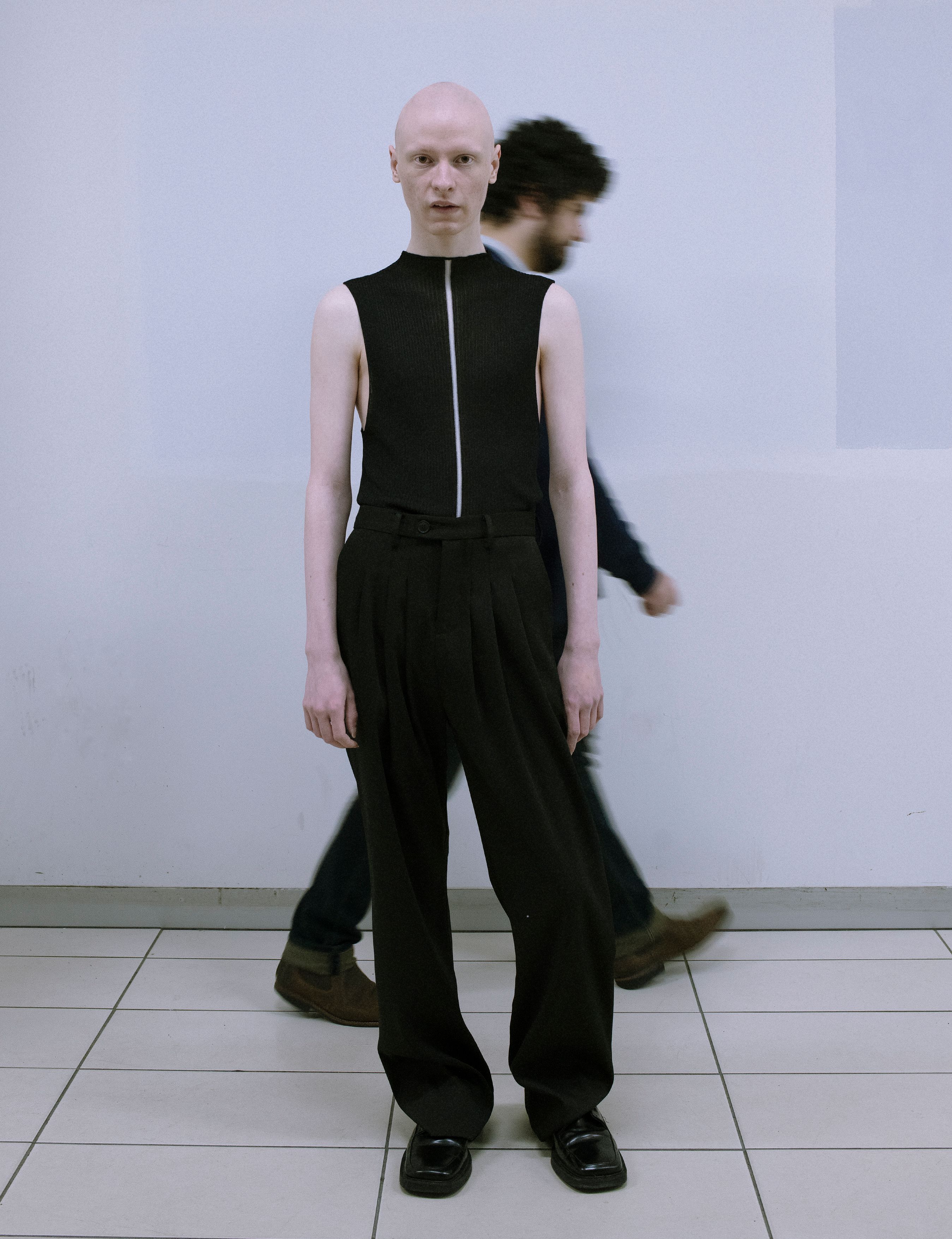 Bald person in black sleeveless top and wide black pants standing indoors with blurred figure in the background, full shot, eye-level.