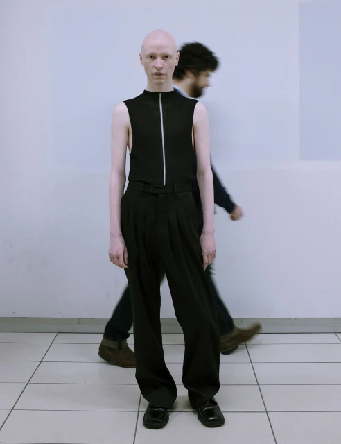 Bald person in black sleeveless top and wide black pants standing indoors with blurred figure in the background, full shot, eye-level.