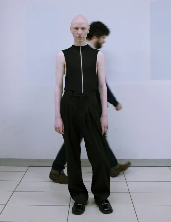 Bald person in black sleeveless top and wide black pants standing indoors with blurred figure in the background, full shot, eye-level.
