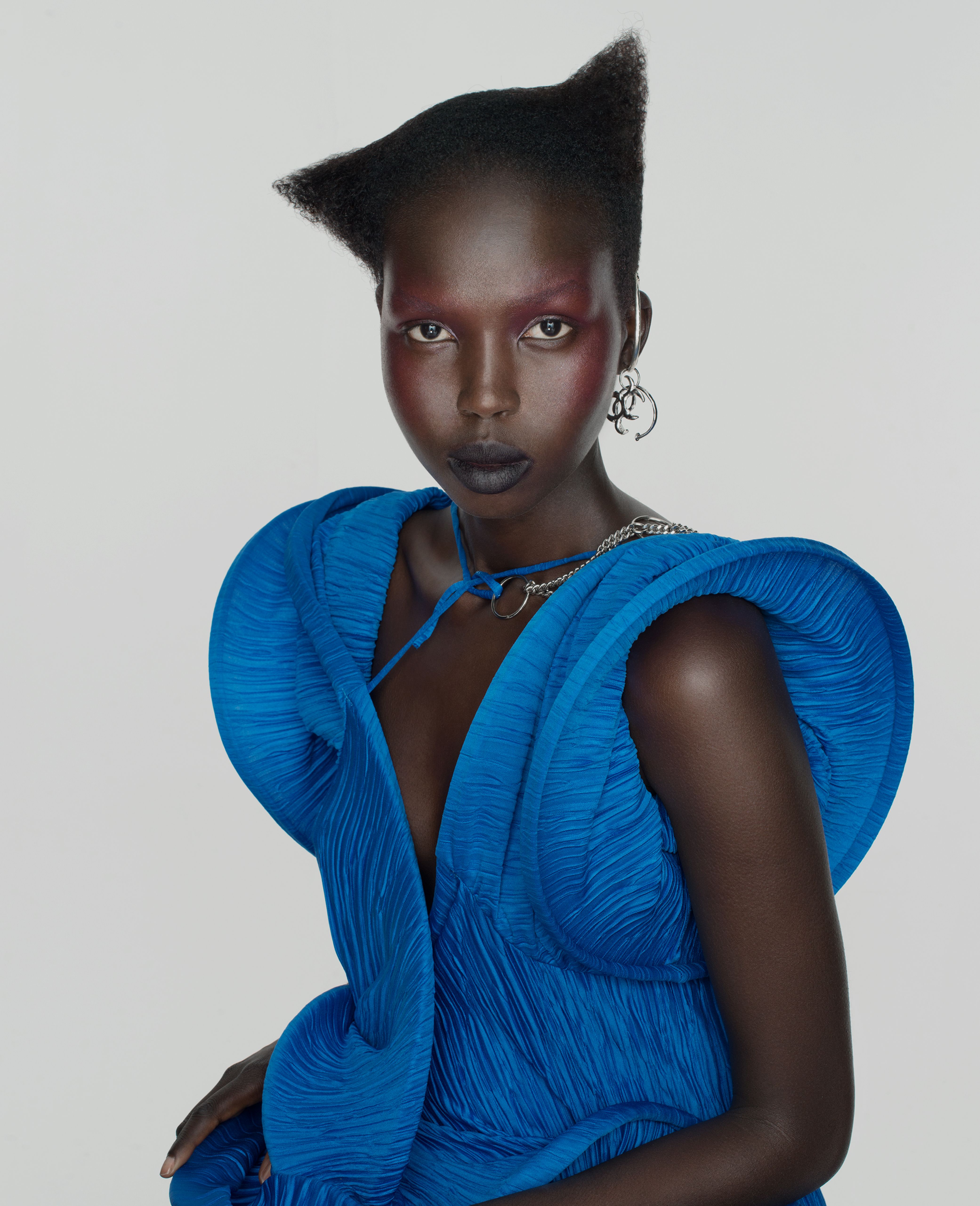 Portrait of a woman with dark skin, wearing a blue textured dress and silver jewelry, against a light gray backdrop in a studio setting.