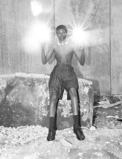 Black and white portrait of a Black male model in shorts and sneakers posing outdoors holding bright lights with a concrete background.