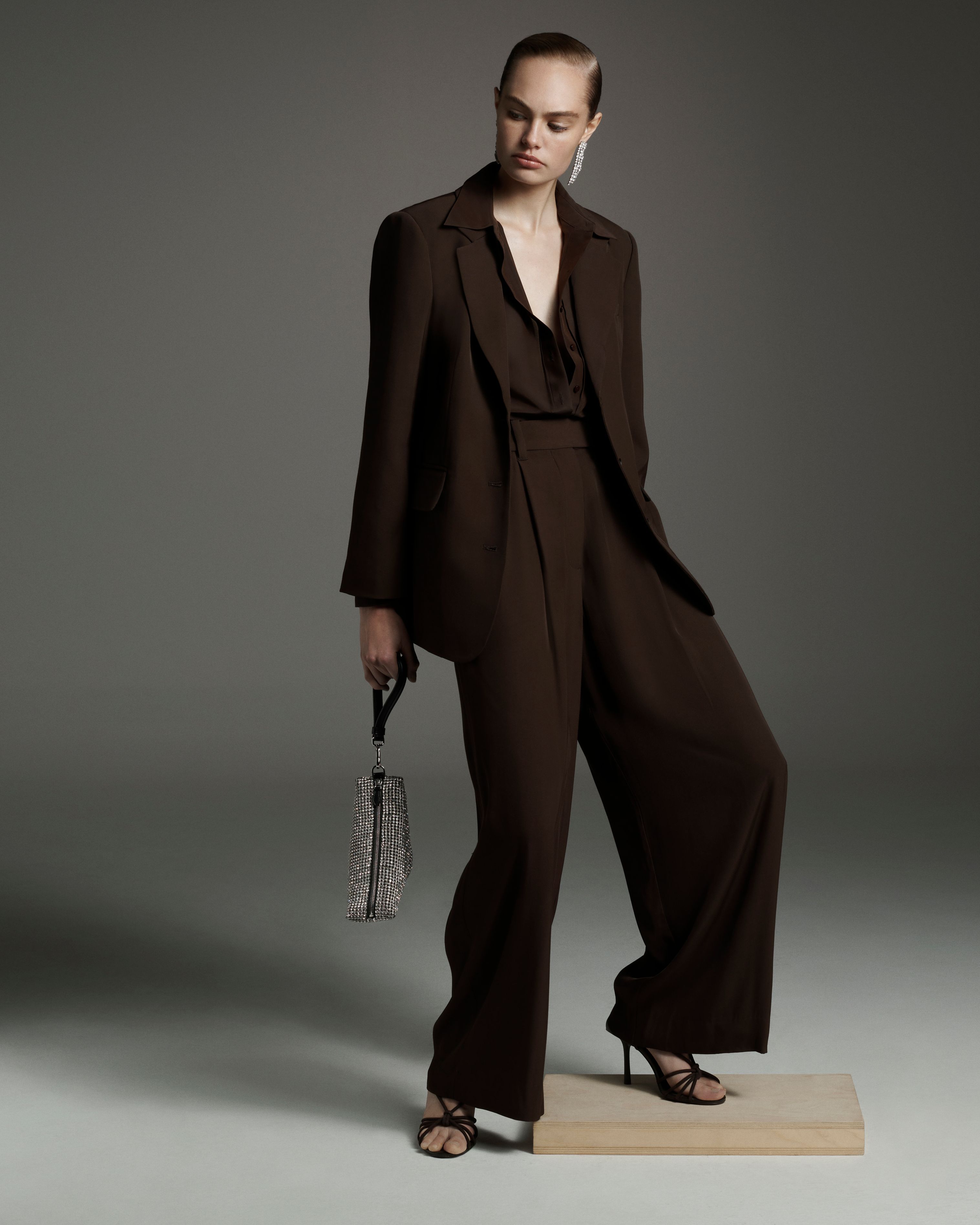Fashion model poses in a brown suit with wide-leg pants, holding a silver purse, standing on a wooden block against a grey background.
