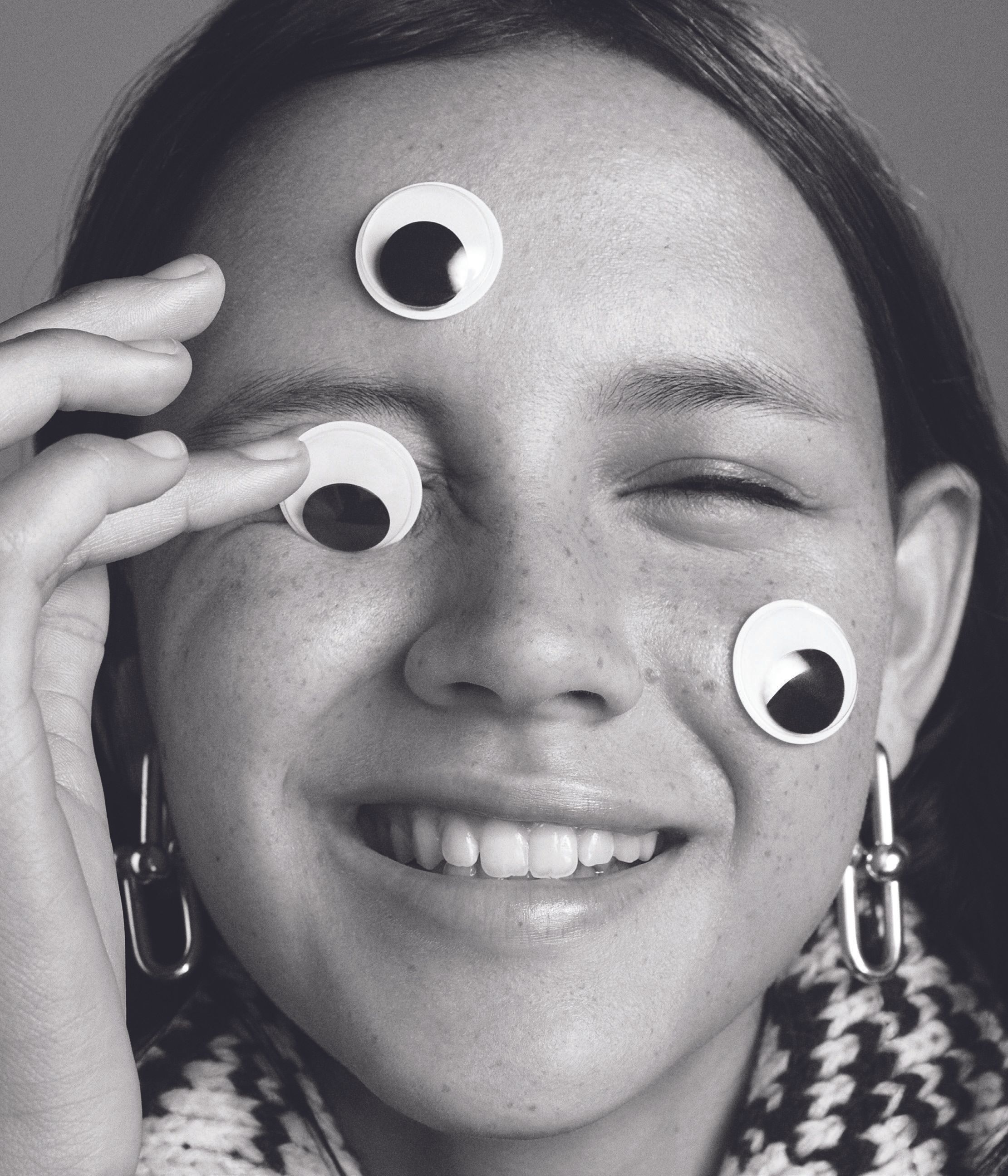 Close-up black and white portrait of a smiling person with googly eyes attached to their face, wearing patterned knitwear and earrings.