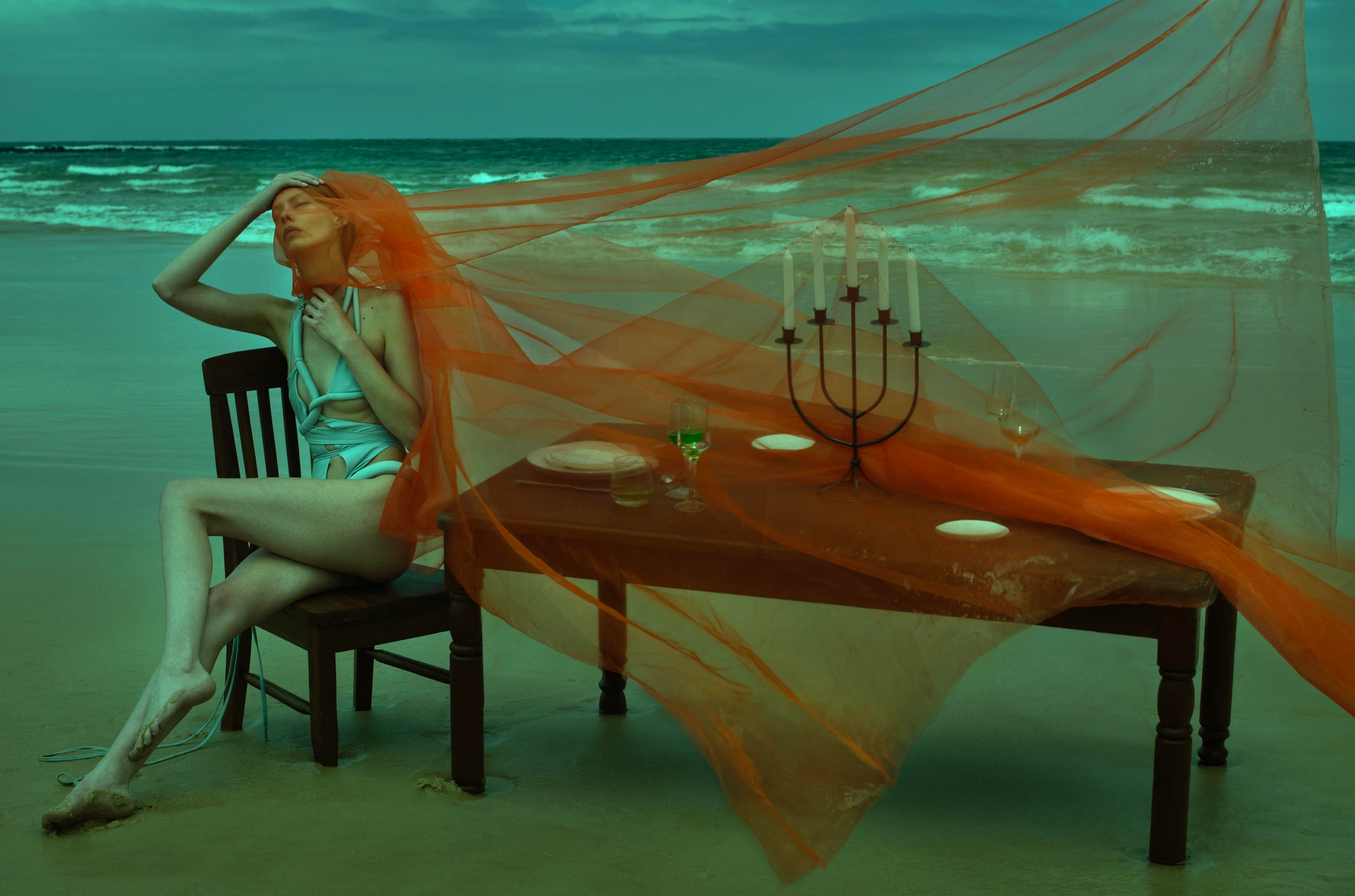 Woman in swimsuit seated at a wooden dining table on a sandy beach draped in orange fabric by Edward Mulvihill