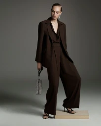 Model in brown pantsuit holding patterned handbag posing on wooden block against grey studio backdrop