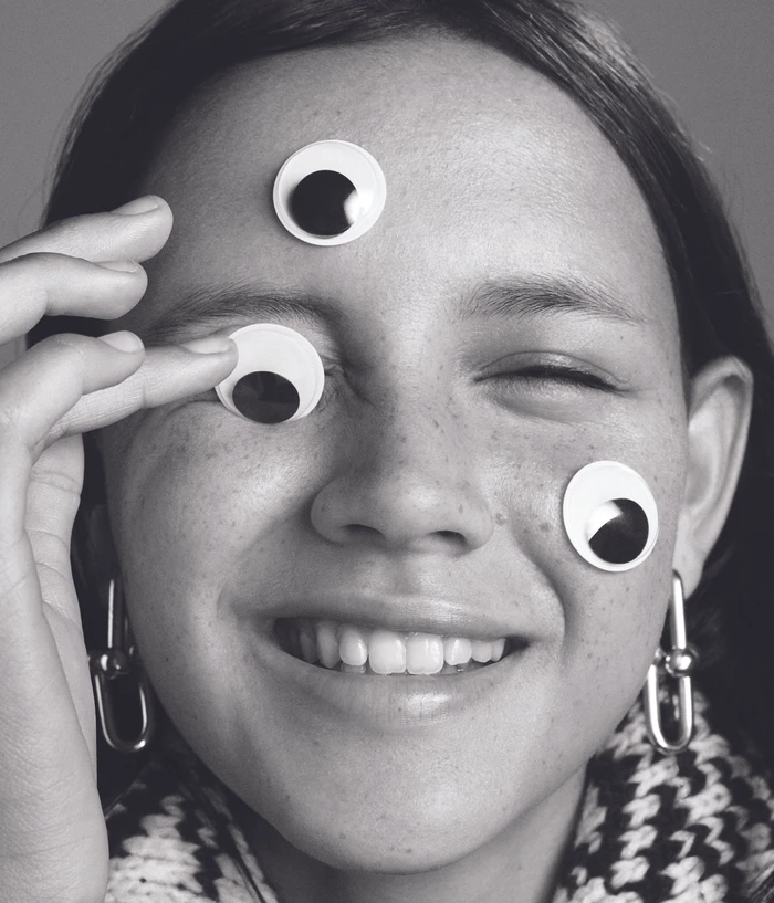 Close-up portrait of a smiling woman with googly eyes on her forehead, cheek, and eyelids photographed by Edward Mulvihill