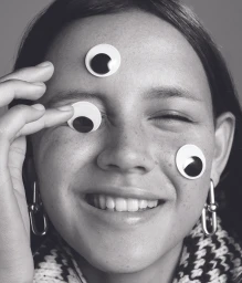Close-up portrait of a smiling woman with googly eyes on her forehead, cheek, and eyelids photographed by Edward Mulvihill