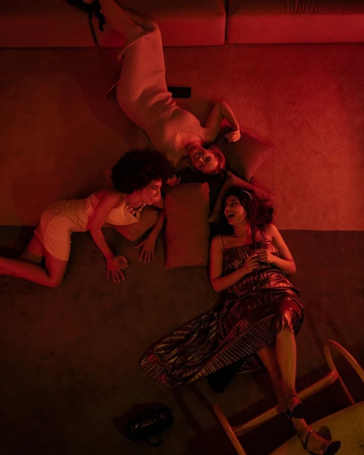 Three women in dresses lounge on floor cushions under red light, talking and laughing in a cozy room, creating a relaxed party scene.
