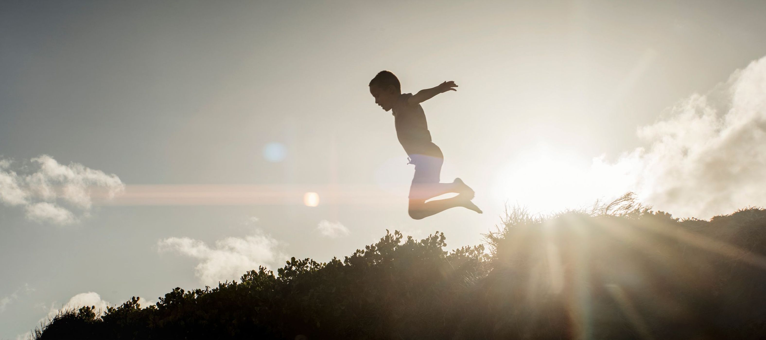 Silhouette of a child jumping mid-air against a bright sunset sky and silhouetted foliage, photographed by Dick Sweeney.